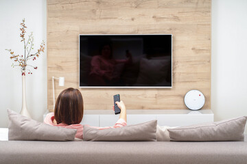 Back view of lonely sad depressive young woman in luxury apartments, sitting on sofa, watching TV...