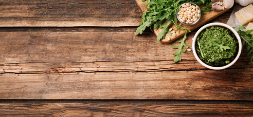 Bowl of tasty arugula pesto and ingredients on wooden table, flat lay with space for text. Banner design