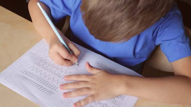 Child boy writing different lines with pencil. Prewriting practice to prepare hands for write letters. Children education concept. View from above