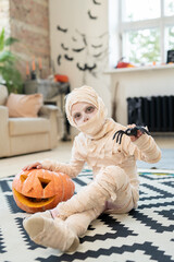 Portrait of mysterious boy dressed up mummy costume sitting on carpet and leaning on jack-o-lantern...