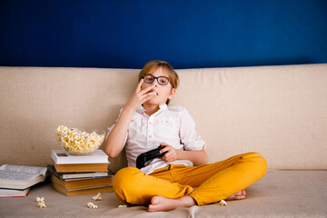 Blonde schoolboy boy plays video games, holds a gamepad, eats popcorn instead of learning lessons...