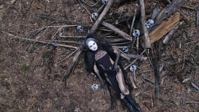 Woman in image of witch lies on ground near fallen trees and human skulls.