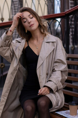 attractive slim young brunette in fashionable trench coat and black dress sitting on a bench with a newspaper. autumn, yellow leaves.