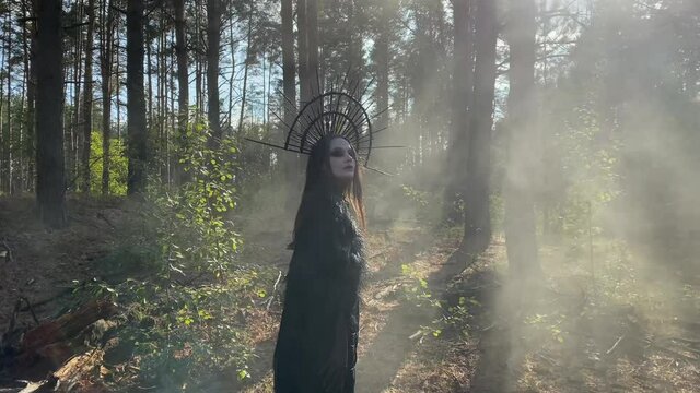 Woman in image of witch stands in penetrated by the sun rays foggy forest.