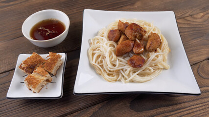Rice noodles with fried chicken pieces in a white square plate next to fried bread and sauce on wooden background