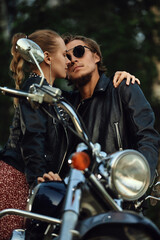 Fototapeta na wymiar young couple of bikers closely looking at each other on black motorcycle on road near green forest