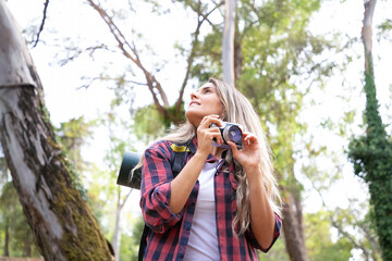 Pretty young woman holding camera and looking around. Happy female traveler hiking with backpack, shooting landshate or taking photo and smiling. Backpacking tourism, adventure and vacation concept