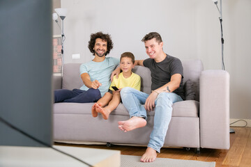 Happy gay fathers and son watching funny TV show at home, sitting on couch in living room, smiling...