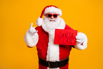 Portrait of his he nice attractive cheerful bad Santa holding in hands red board placard showing horn symbol party hard isolated over bright vivid shine vibrant yellow color background