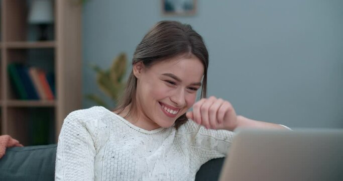 Young woman having video conversation while using laptop. Pretty female person smiling and telling with sign language I love you while sitting on sofa at home. Concept of hearing loss.