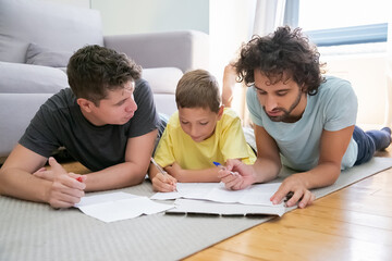 Couple of homosexual parents helping focused boy with school home task, lying on floor at home,...
