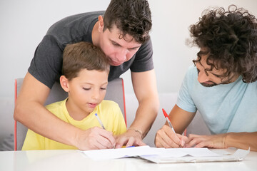 Fototapeta premium Gay parents helping focused boy with school home task, sitting at table and writing in papers together. Family and parenthood concept