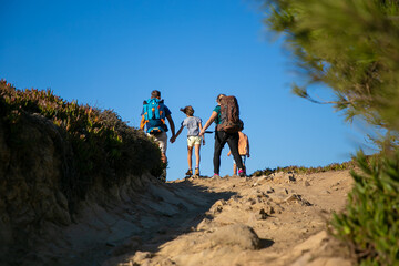 Family of hikers with backpacks walking on track. Parents and two kids hiking outdoors. Back view. Hiking or adventure tourism concept