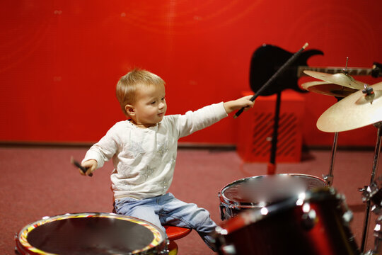 Toddler Kid Playing Drums On Red Background, Kid Playing Drum Kit, Concept Musical Development From Early Childhood