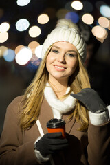 beautiful girl, in the evening, against the background of lights with a drink in an orange glass, a white hat and a beige coat. smiling at camera