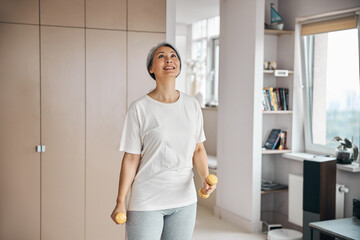 Fototapeta na wymiar Adult Asian female in white shirt holding sport tools in hands while looking up in living room