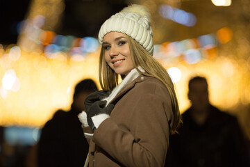 beautiful girl, in the evening, against the background of lights, a white hat and a beige coat. smiling at camera