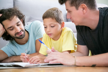 Fototapeta premium Two fathers helping focused boy with school home task, lying on floor at home, writing or drawing in papers. Family and gay parents concept