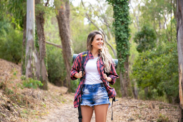 Happy woman standing on forest road, smiling and looking away. Long-haired woman carrying backpacks and hiking on nature. Green trees on background. Tourism, adventure and summer vacation concept