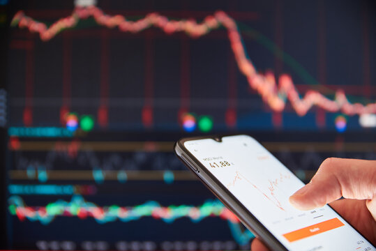 Investor Analyzing Stock Market Investments On A Smartphone. Person Trading Stocks On A Smartphone In Front Of A Big Screen With Falling Share Prices. Stock Market Crash. US Stock Exchange.