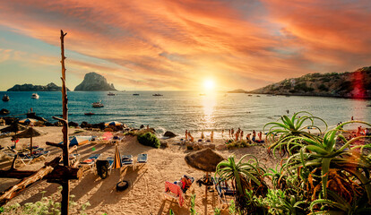 Picturesque view of Cala d'Hort tropical Beach, people hangout in beautiful beach with Es Vedra rock view during magnificent vibrant sunset glowing sun. Balearic Islands, Spain, Espana. Ibiza - obrazy, fototapety, plakaty