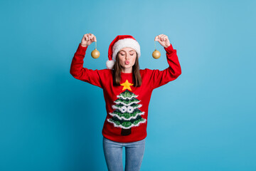 Portrait photo of girl wearing xmas jumper and headwear keeping showing decorative golden christmas balls baubles with plump pouted lips sending air kiss isolated on blue color background