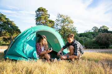 Caucasian young tourists camping on lawn and sitting in tent. Happy couple drinking tea from thermos and relaxing on nature together. Backpacking tourism, adventure and summer vacation concept