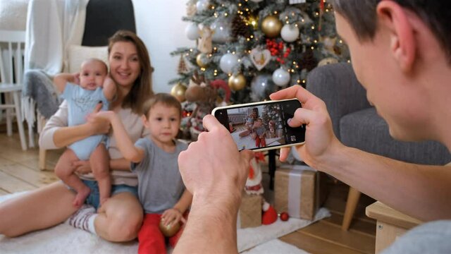 Father Making Family Photos by Phone at Christmas. Young Woman with Little Baby and Older Son Posing Near Xmas Tree. Selective Focus. Christmas, New Year, Winter Holiday Celebration