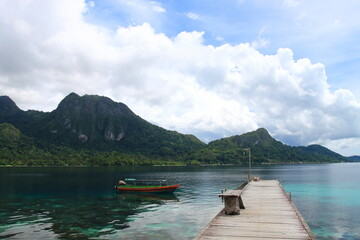 Obraz premium The wooden pier over the sea with green hills and cloudy sky on the background at Ora Beach Resort, Central Maluku, Indonesia