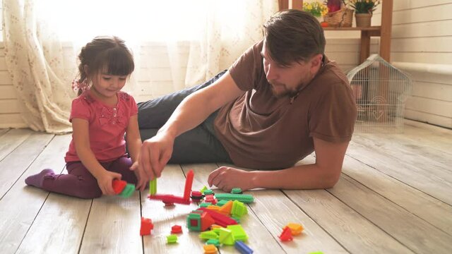 Little beautiful girl and her dad are sitting on the floor in the room and playing with a colorful constructor
