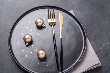 Christmas table setting. Black ceramic plate with golden balls and cutlery on stone background. Gold decoration