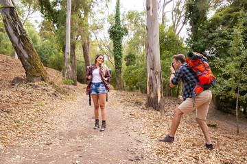 Blonde woman posing for photo on road in forest. Caucasian man holding camera and shooting on nature. Two happy people trekking with backpacks. Tourism, adventure and summer vacation concept