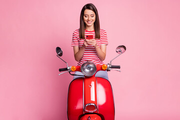 Photo portrait of surprised woman sitting on red moped texting on smartphone smiling isolated on pastel pink color background