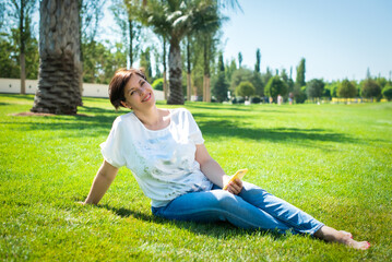 pretty middle-aged woman sits on the grass under palm trees with a smartphone. work online in...