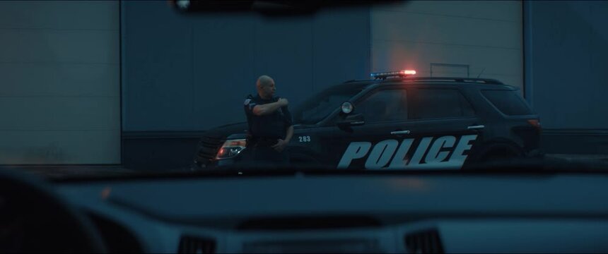 Caucasian male police officer standing guard in the street near police car with flashing lights, talking on CB radio. Shot on RED cinema camera with 2x Anamorphic lens