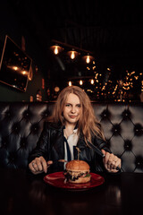Portrait of a young lovely hungry woman sitting in the street fast junk food restaurant cafe hold fork and knife and eating enjoying a fresh tasty fat burger on a red plate. Image with copy space.
