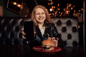 Portrait of a young lovely hungry woman sitting in the street fast junk food restaurant cafe hold showing smartphone mock up screen and eating enjoying a fresh tasty burger on a red plate.
