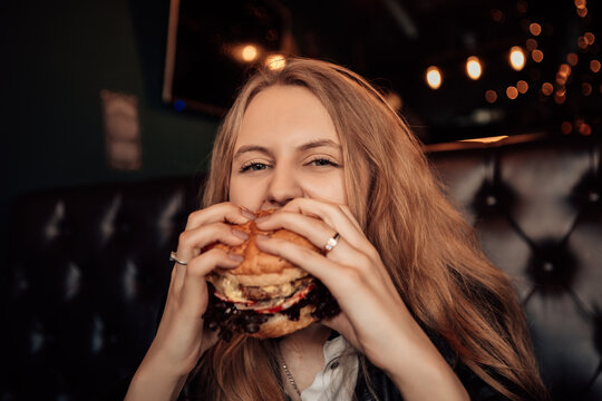 Portrait of a young lovely hungry woman sitting in the street fast junk food restaurant cafe with open mouth bite and eating enjoying a fresh tasty burger. Image with copy space.

