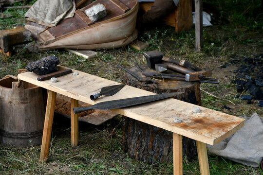 A close up on two metal weapon elements laying on a wooden bench next to some blacksmith equipment and a bucket of water seen on a sunny summer day during a traditional medieval fair in Poland
