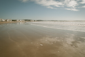 Empty beach at the Atlantic coastline. Smooth sand washed by the sea. Clouds reflected in water. Small maritime town in the distance. 