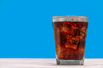 Ice cold glass of soda, cola, fizzy drink, carbonated beverage sits against a blue wall with ice...