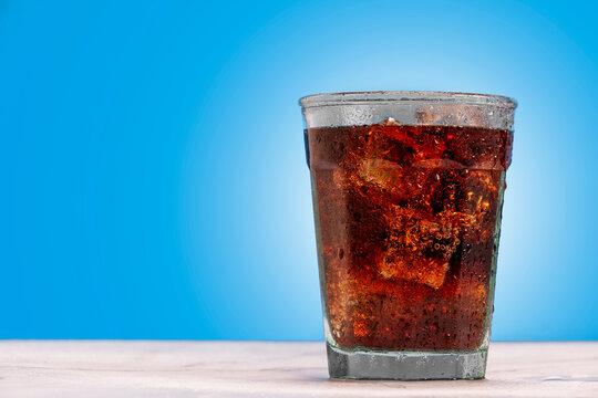 Ice cold glass of soda, cola, fizzy drink, carbonated beverage sits against a blue wall with ice cubes and condensation on the glass making it refreshing and inviting.
