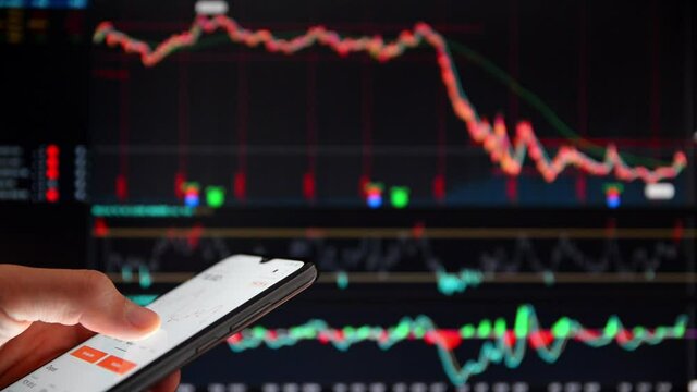 Investor analyzing the stock prices on a smartphone. Person with smartphone in front of a big screen with falling share prices. Dropping stock prices on a screen. Falling market rates on a screen.