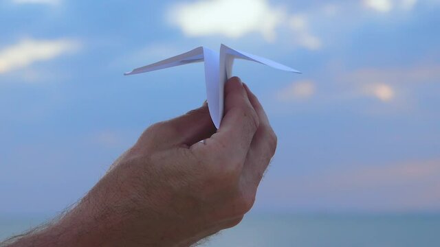 A man's hand holds a paper airplane in close-up against the sky.