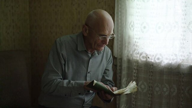 elderly bald man wearing eyeglasses reading old book with antique pages and looking at window indoor