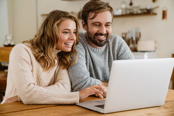 Beautiful happy couple using laptop while sitting in kitchen