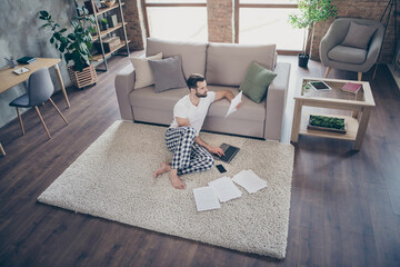 Nice attractive focused guy sitting on carpet studying learning working remotely online analyzing documents at modern loft industrial open space interior style flat indoors