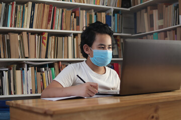 Asian boy student wearing protective mask learning studying online with laptop computer in library, school from home