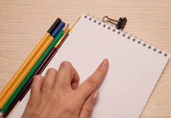 Painting brushes, pencil, pens, paper clip, female left hand finger pointing at white blank notebook on wooden background