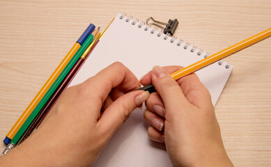 Drawing brushes, pencil, pens, paper clip, white blank notebook and both female hands open the pen on a wooden background
